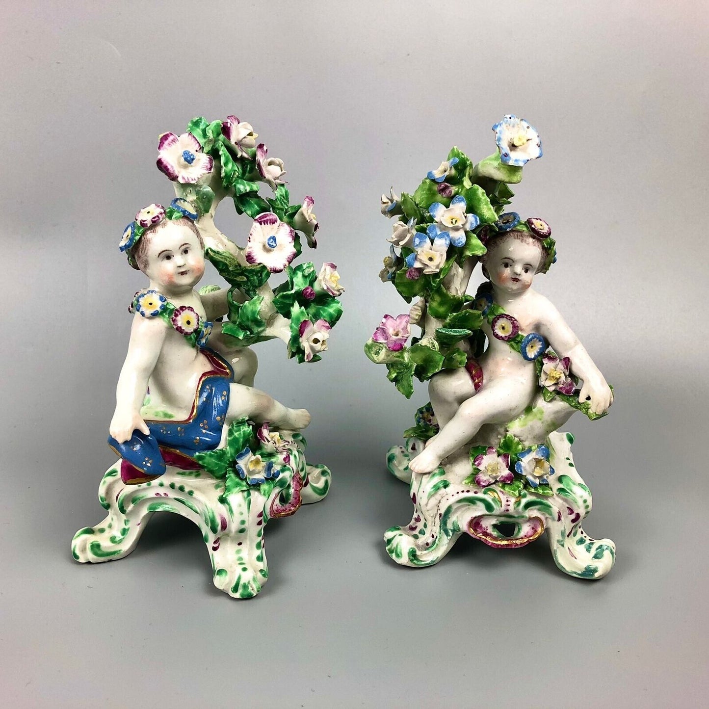 A pair of Bow putti with rococo C-scroll bases and bocage c.1760
